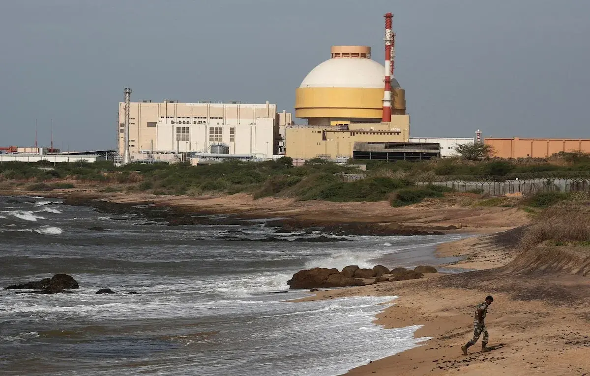 A domed reactor building behind a rocky shoreline, with a policeman walking on the beach